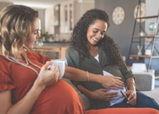 Two pregnant women socialising over a hot beverage. One mother is holding her baby bump and another mother holds her hand out
