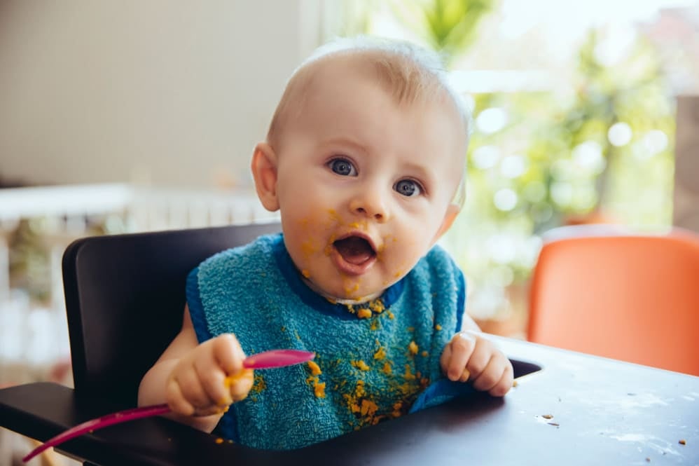 Child sitting in high chair feeding themself with a spoon, with food spilled on the face, bib and tray during mealtime