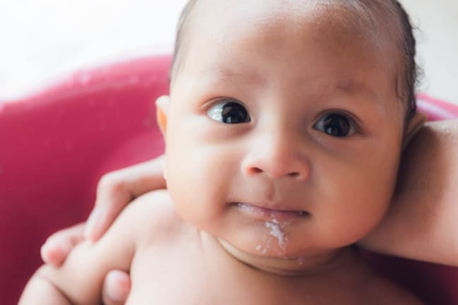 Close-up of a baby being held by an adult, with a small amount of milk spit-up visible around the mouth and chin.